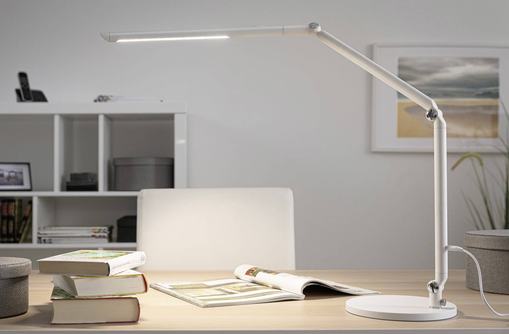 Ein moderner Schreibtisch mit einer weißen LED-Lampe, geöffneten Büchern und Regalen im Hintergrund. Ein Bild an der Wand.