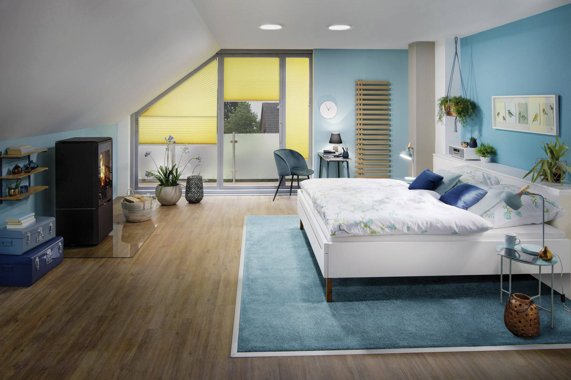 Helles Schlafzimmer mit blauem und gelbem Dekor, großem Bett, Sessel am Fenster, Kamin, Pflanzen und blauen Teppichen auf Holzfußboden.