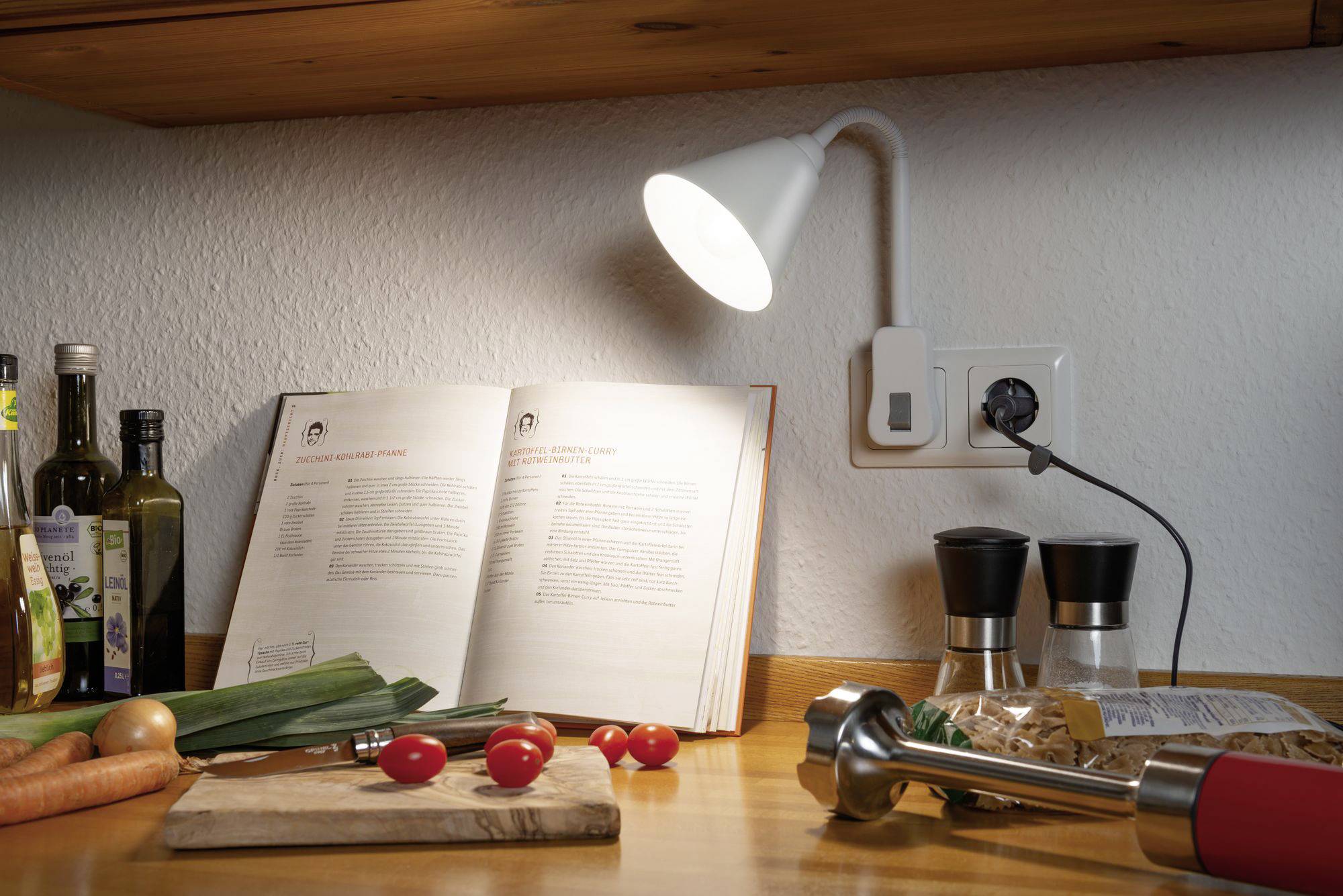 Auf einer Küchenarbeitsplatte liegt ein geöffnetes Kochbuch unter einer Lampe. Daneben sind Gewürze, Tomaten, Karotten und ein Mixer.