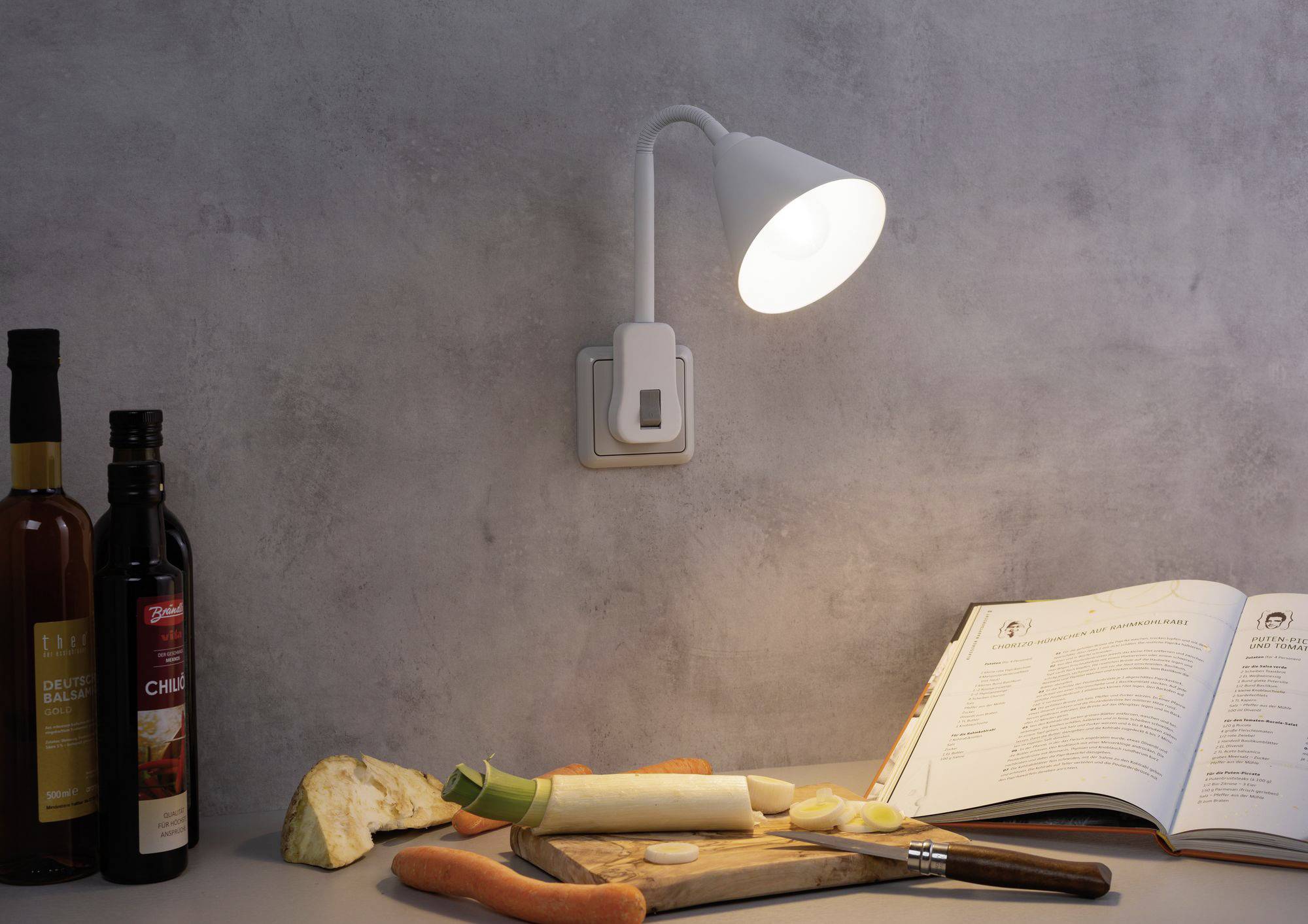 Ein beleuchtetes Rezeptbuch auf einem Tisch mit geschnittenem Gemüse, Messer, Holzbrett und Weinflaschen. Hintergrund: graue Wand.