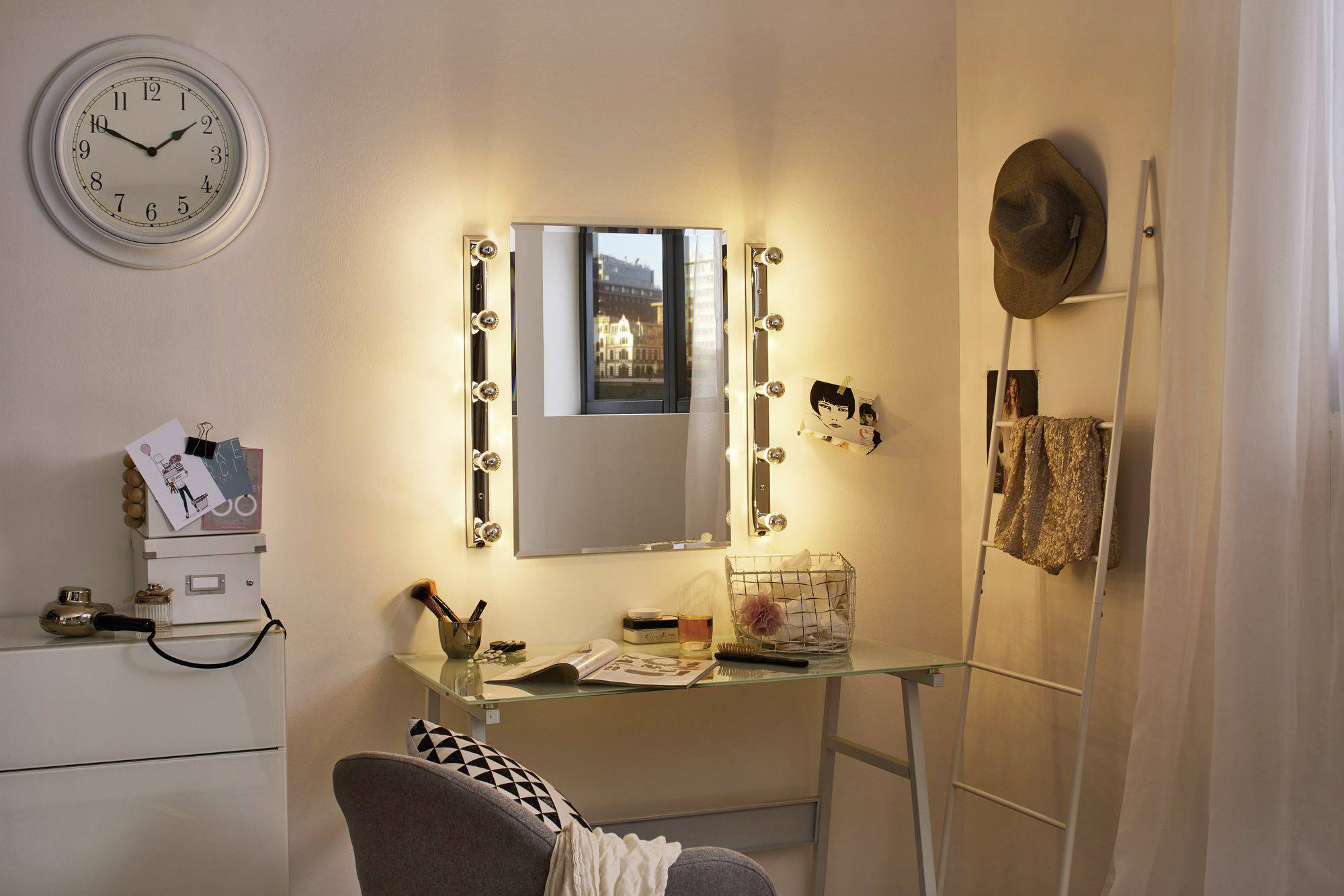 Ein gemütlicher Schminktisch in einem Schlafzimmer, beleuchtet von kleinen Glühbirnen um einen rechteckigen Spiegel, daneben eine Uhr an der Wand.