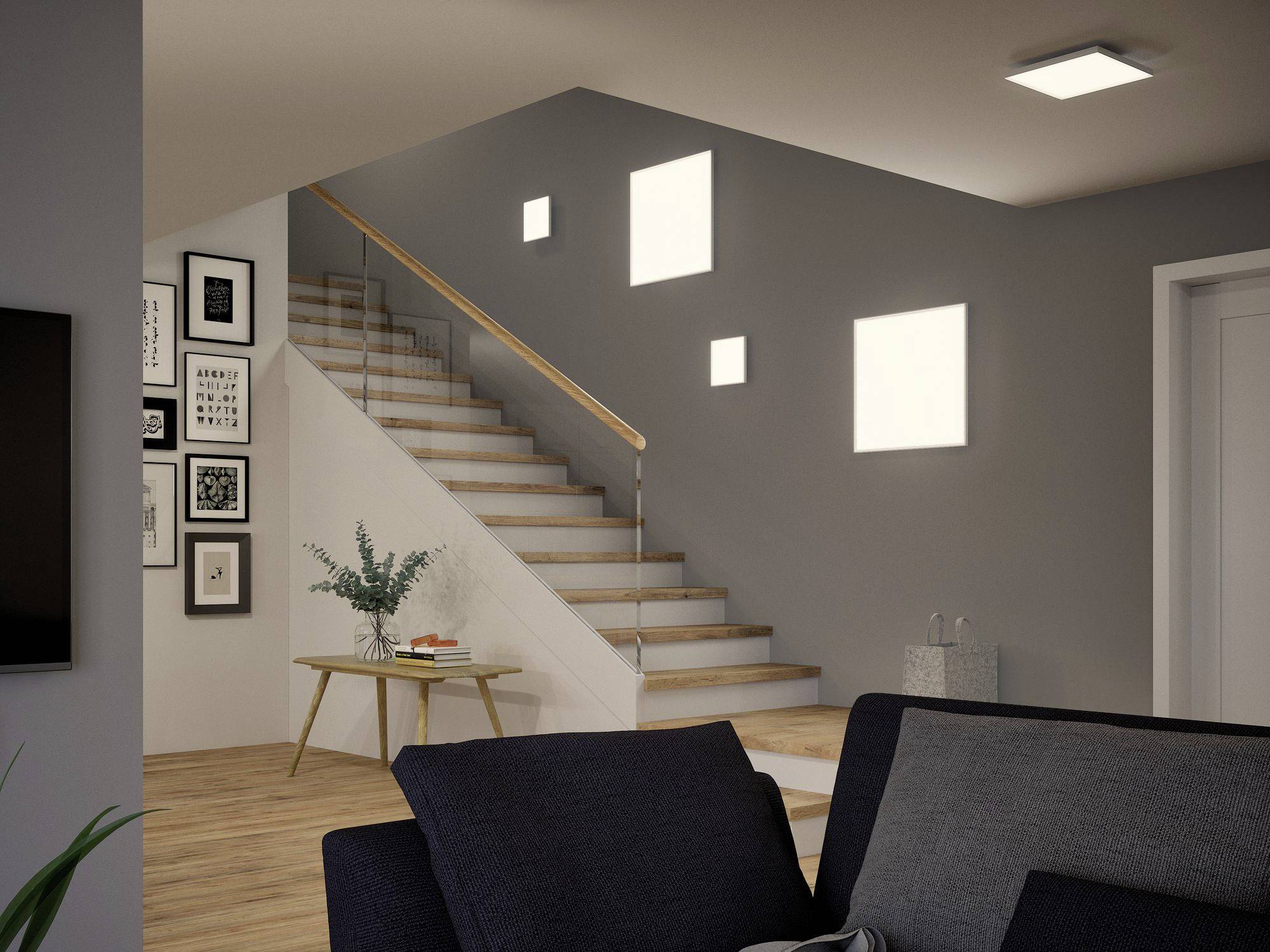 Moderne Wohnzimmer mit Treppe. An der Wand hängen Bilderrahmen, ein Tisch mit Pflanzen steht daneben. Helle Beleuchtung betont den Raum.
