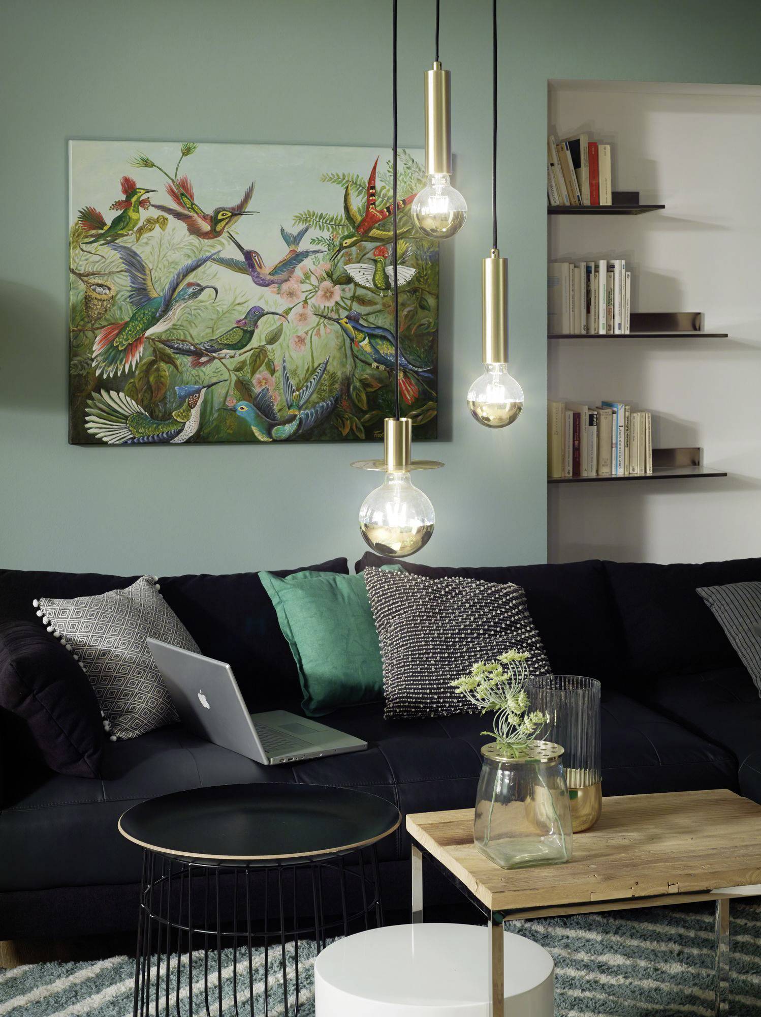 Ein gemütliches Wohnzimmer mit einem schwarzen Sofa, einem Laptop auf einem runden Tisch und einem bunten Vogelgemälde an der Wand.