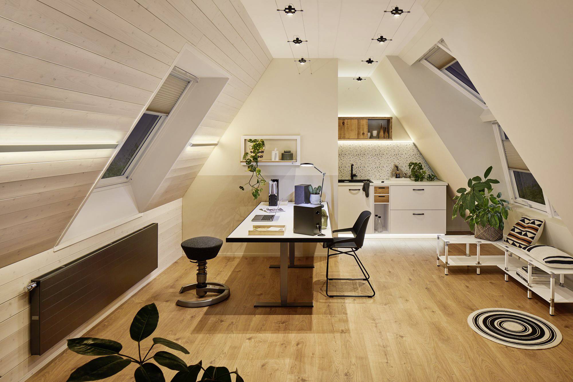 Modernes Dachgeschoss-Büro mit Schreibtisch, Laptop, Pflanzen und kleiner Küche. Warmes Holzinterieur und Schrägdachfenster.