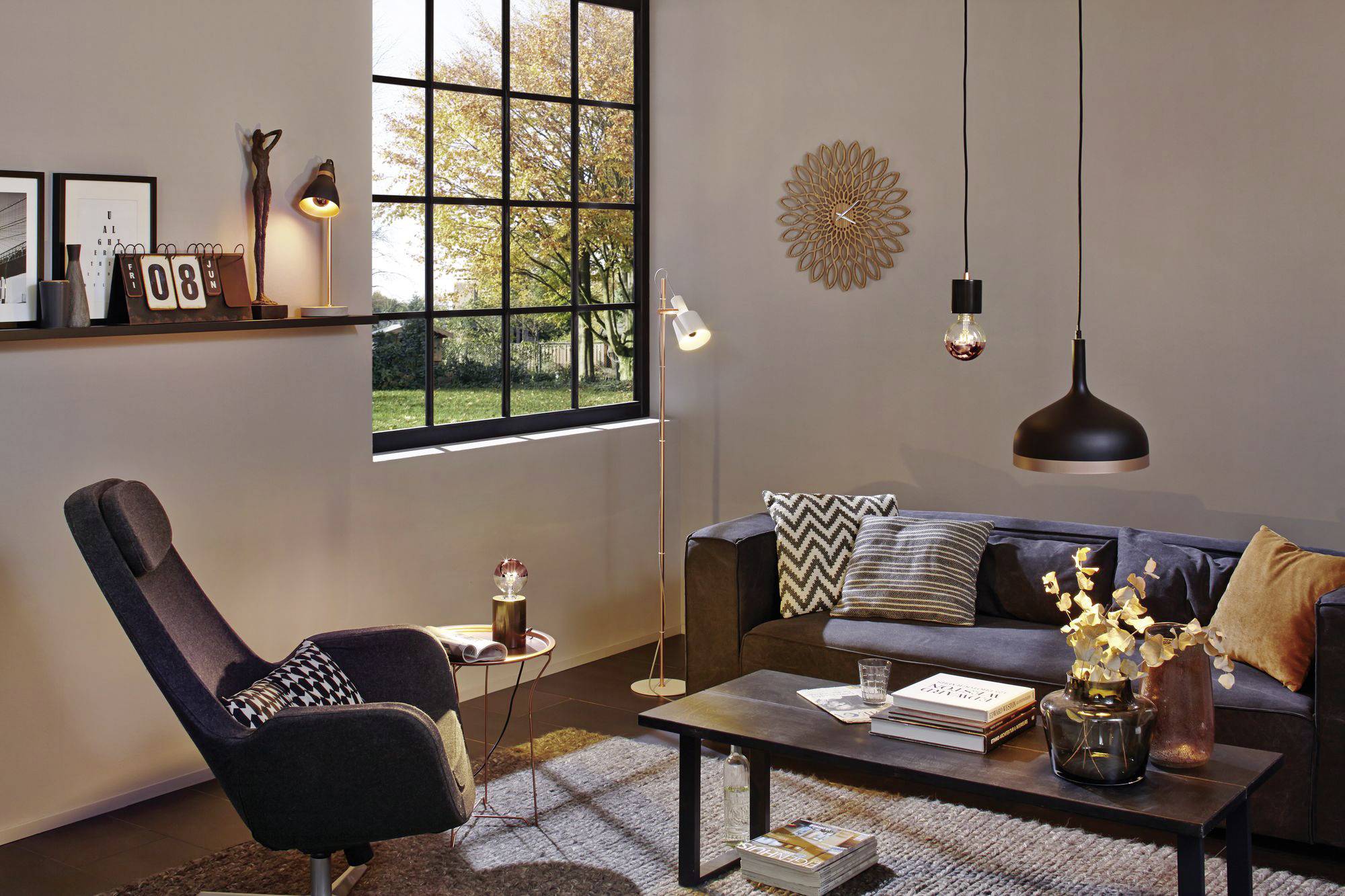 Wohnzimmer mit Sofa, Leselampe, Sessel und Couchtisch. Große Fenster bieten Ausblick auf einen Baum im Herbst. Moderne, gemütliche Einrichtung.
