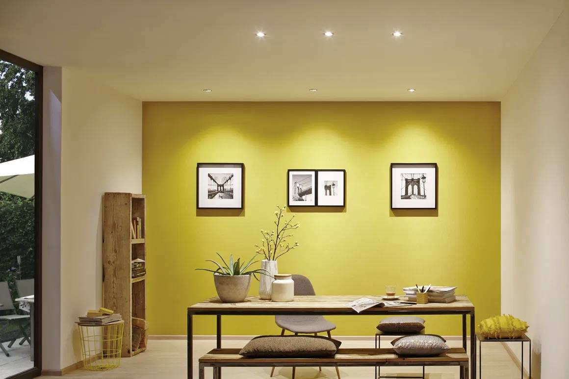 Ein modern eingerichtetes Esszimmer mit gelber Akzentwand. An der Wand hängen vier gerahmte Bilder. Ein Tisch mit Stühlen und Pflanzen.