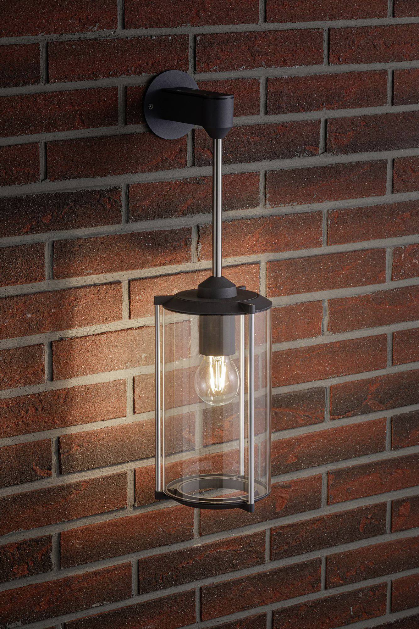 Moderne, schwarze Außenwandleuchte mit klarer Glasscheibe und sichtbarer Glühbirne, montiert an einer rotbraunen Klinkerwand.
