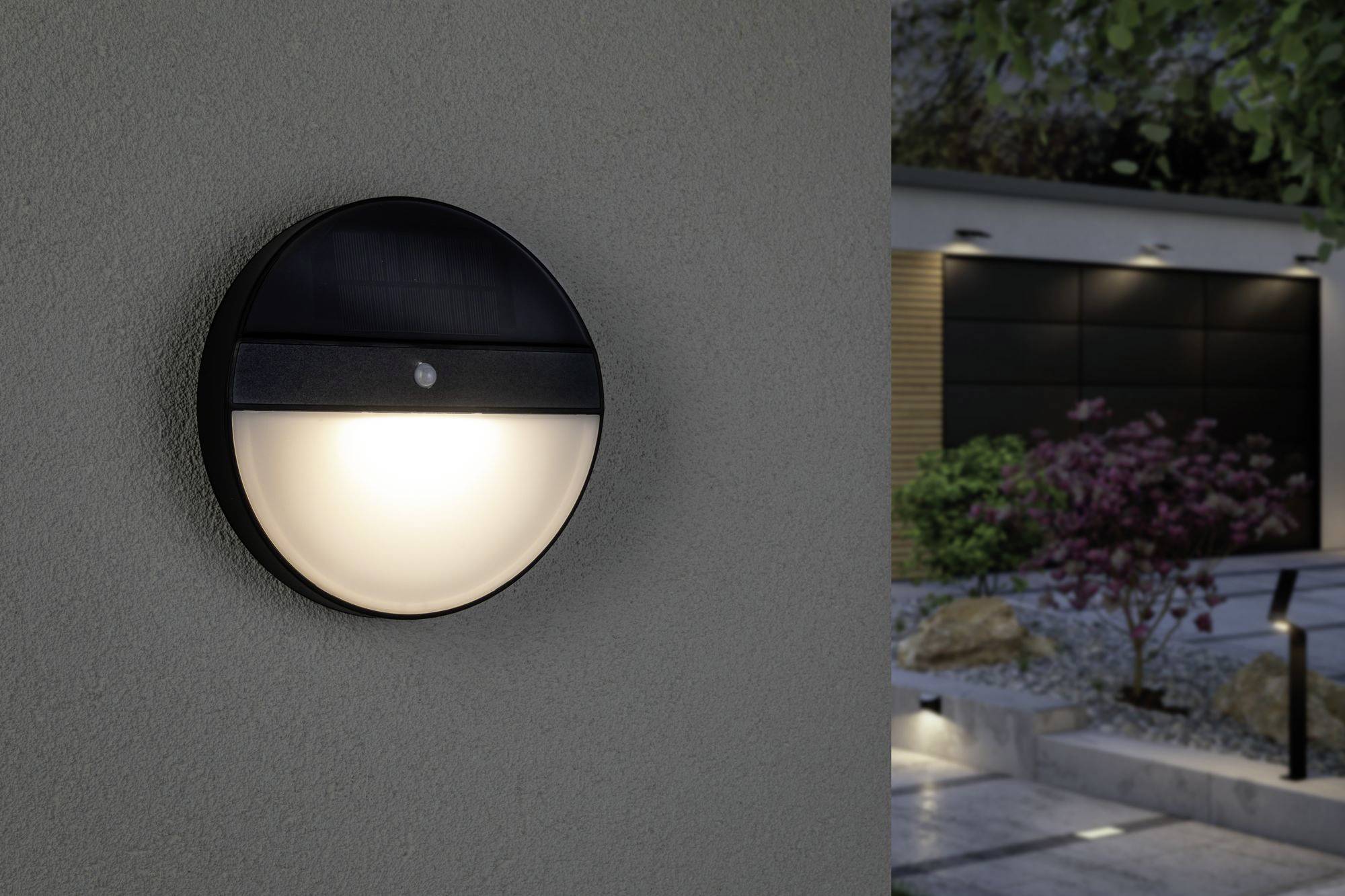 Eine runde Außenleuchte an einer Wand leuchtet. Im Hintergrund ist ein moderner Garten mit Beleuchtung und Pflanzen zu sehen.