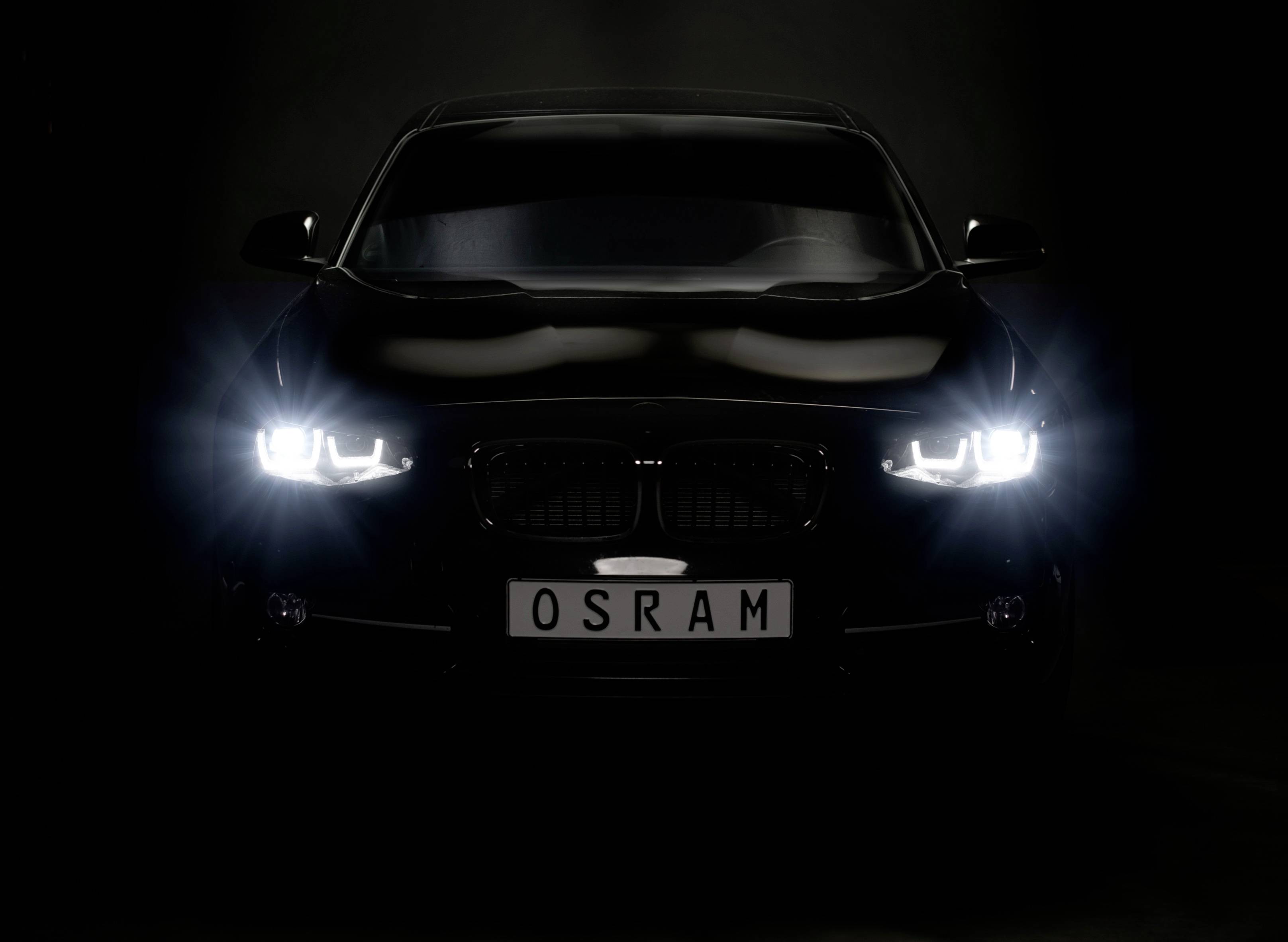Ein schwarzes Auto mit leuchtenden Scheinwerfern steht vor einem dunklen Hintergrund. Das Kennzeichen zeigt 'OSRAM'.