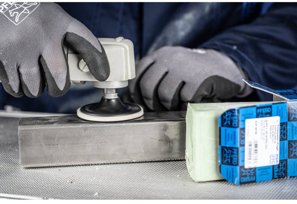 Person mit Handschuhen poliert Metallblock mit Schleifmaschine, daneben grüne Schleifpaste.