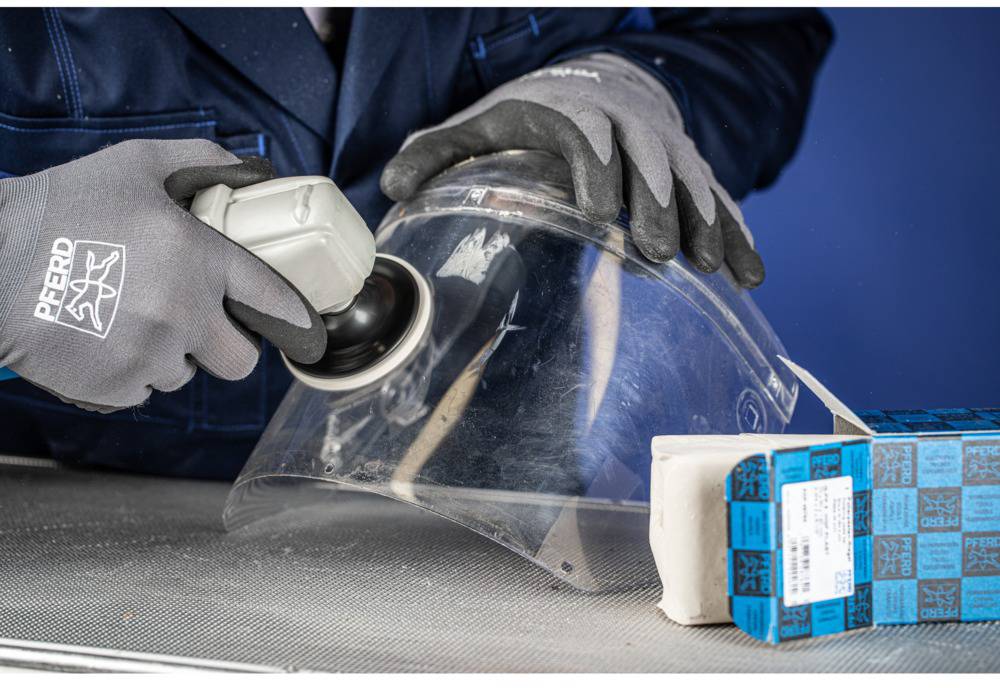 Person schleift transparente Kunststoffscheibe mit Elektrowerkzeug; trägt graue Handschuhe mit Logo, daneben blaue Verpackungen.