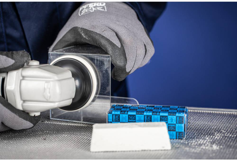 Eine Person schleift eine Metalloberfläche mit einem Winkelschleifer. Der Schleifer hat ein graues Gehäuse, daneben liegt ein blauer Schleifblock.