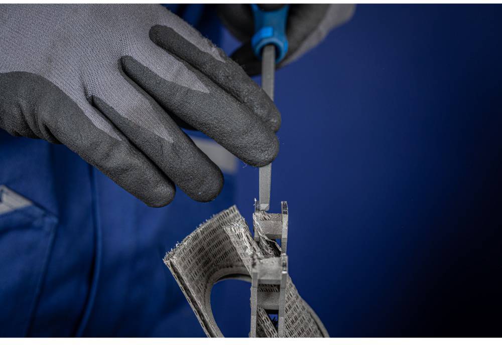 Eine Person in blauer Arbeitskleidung und grauen Handschuhen feilt an einem Metallwerkstück.