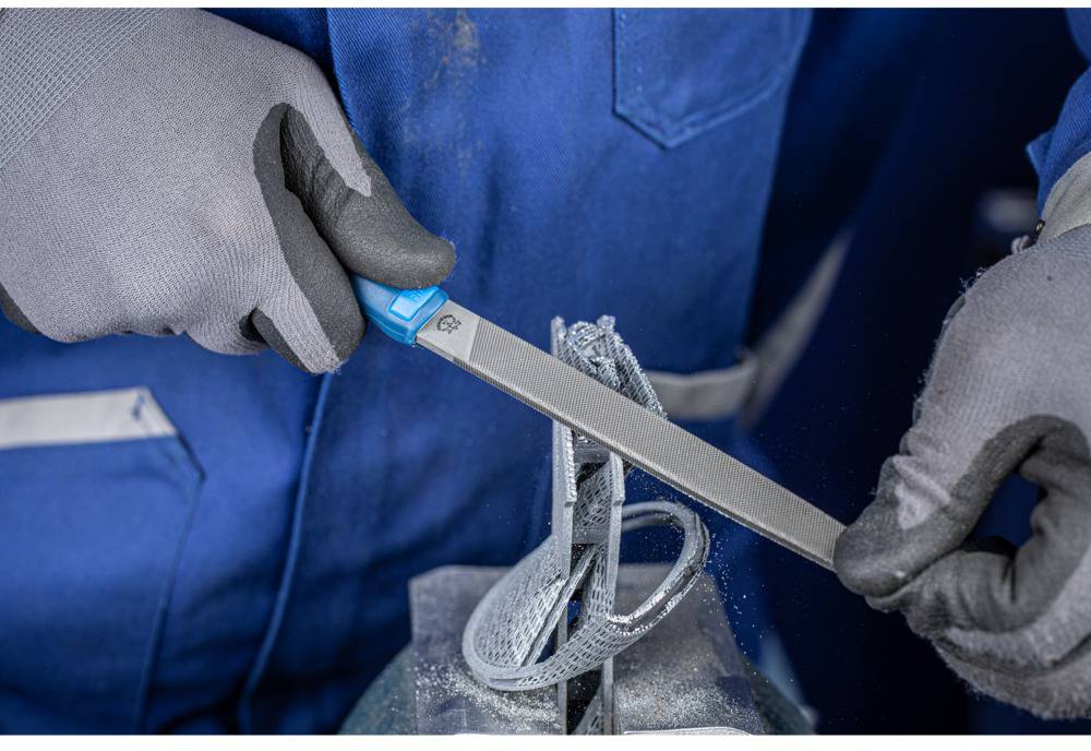 Eine Person in blauer Arbeitskleidung feilt ein metallisches Objekt mit einer Handfeile, umgeben von Metallstaub.