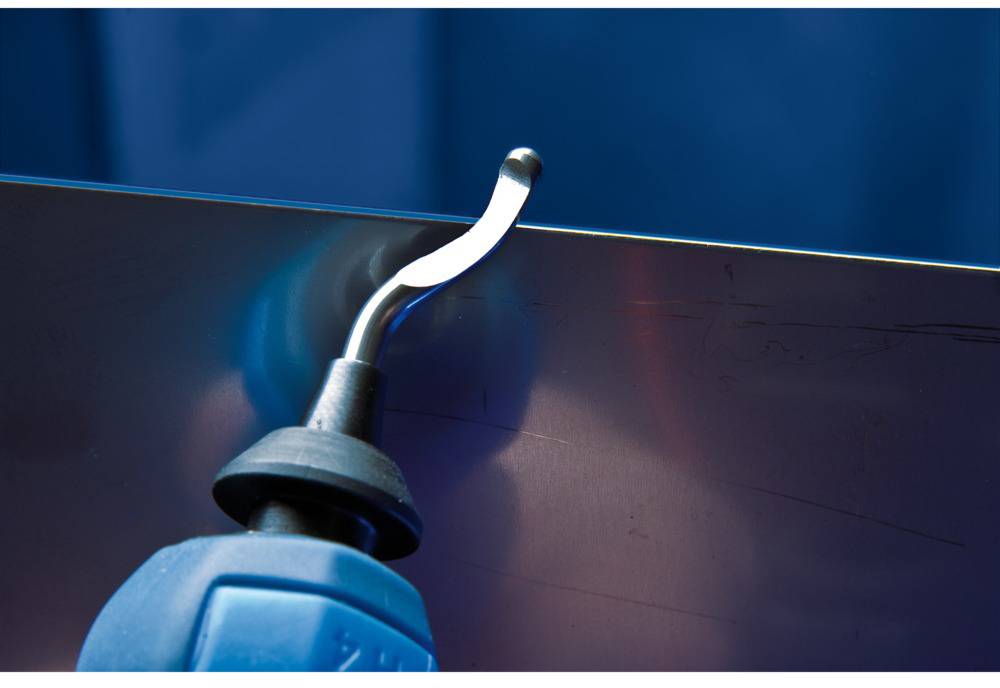Ein Entgratwerkzeug entfernt Kante von einem Metallblech. Der Hintergrund ist unscharf, der Fokus liegt auf dem Werkzeug und dem Blech.