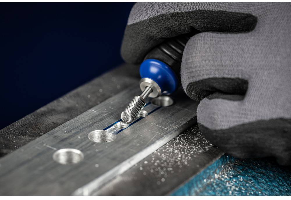Handschuhe tragen ein Rotationswerkzeug über eine Metalloberfläche, um Material zu entfernen. Metallspäne sind erkennbar.