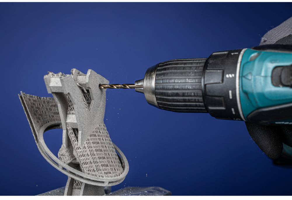 Eine elektrische Bohrmaschine bearbeitet ein 3D-gedrucktes Objekt aus grauem Kunststoff vor blauem Hintergrund.