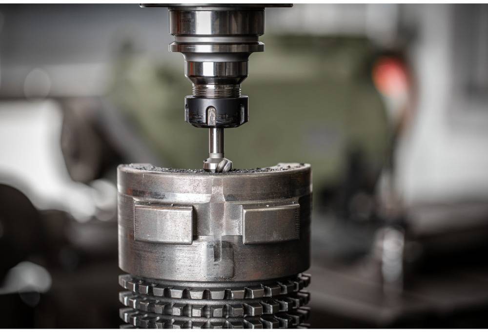 Eine Maschine fräst präzise ein Metallwerkstück. Im Hintergrund ist unscharf ein weiteres Maschinenteil zu sehen.