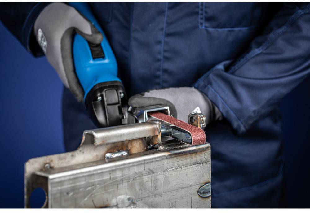 Eine Person in Arbeitskleidung schleift mit einem blauen Winkelschleifer ein Metallstück. Sie trägt Handschuhe für Sicherheit.