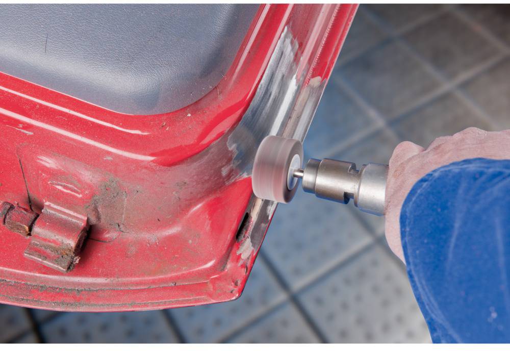 Eine Person verwendet einen Bohrer, um Farbe von einem roten Autoteil zu entfernen. Die Arbeit erfolgt auf einem gefliesten Boden.