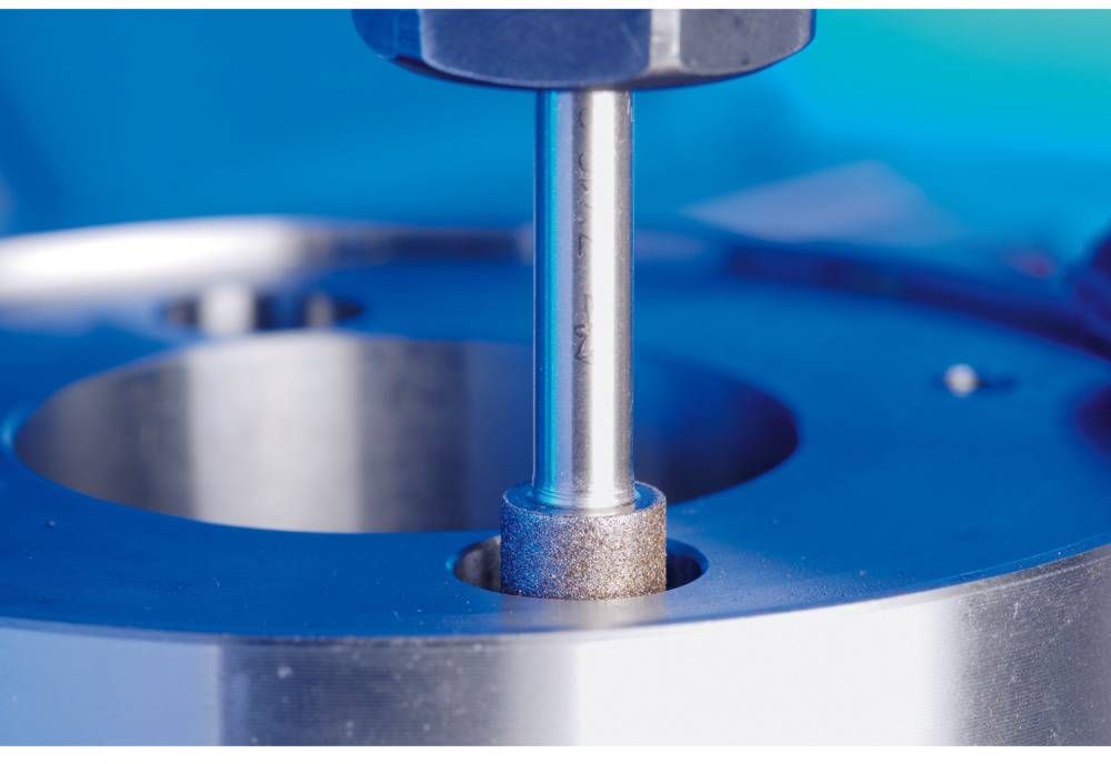 Maschine mit Bohrwerkzeug in Aktion. Fokus auf Zylinder, der in Metallvorrichtung eintritt. Hintergrund in Blau.