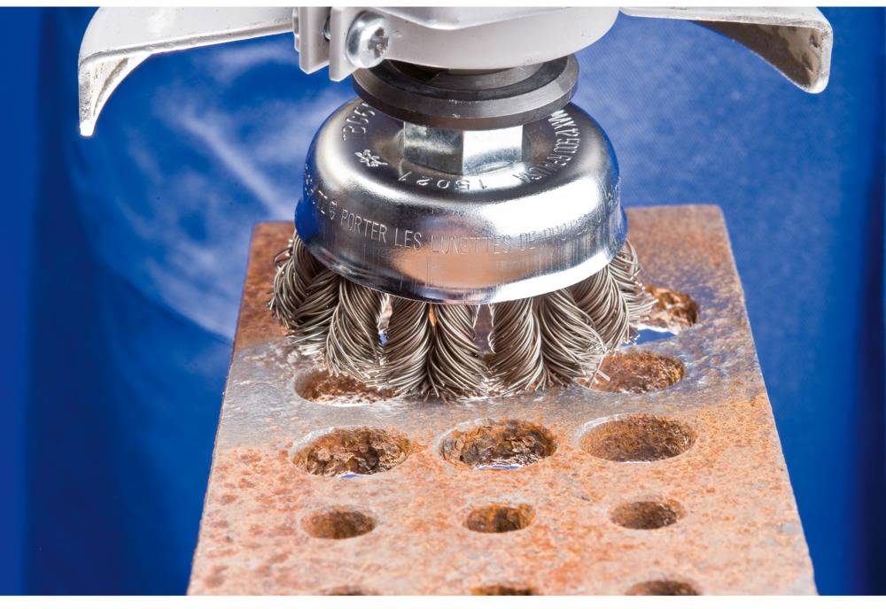 Eine Drahtbürste auf einer Bohrmaschine reinigt eine rostige Metallplatte mit Löchern vor einem blauen Hintergrund.