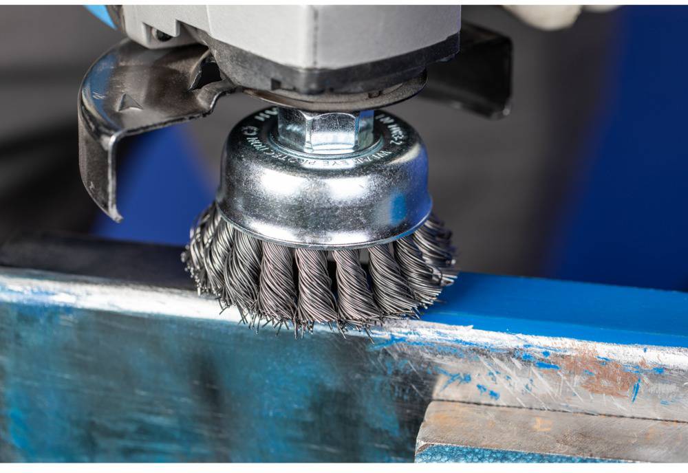 Eine Drahtbürste in Bewegung bearbeitet die Oberfläche eines blauen Metallstücks und entfernt Rost oder alten Lack.