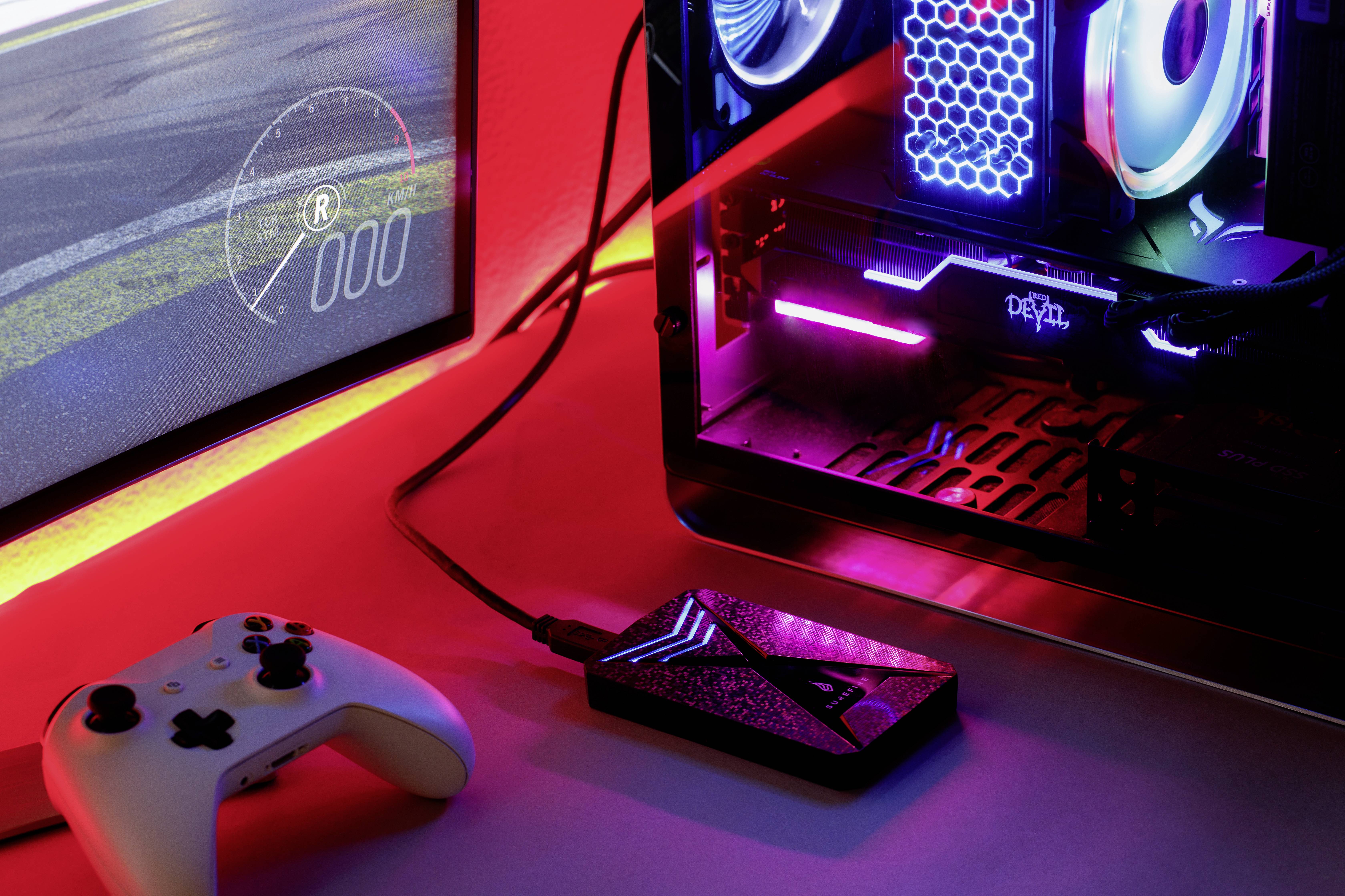 Ein Gaming-Setup mit leuchtendem Desktop-PC, angeschlossenem Gamecontroller und USB-Gerät. Bildschirm zeigt ein Autorennspiel.