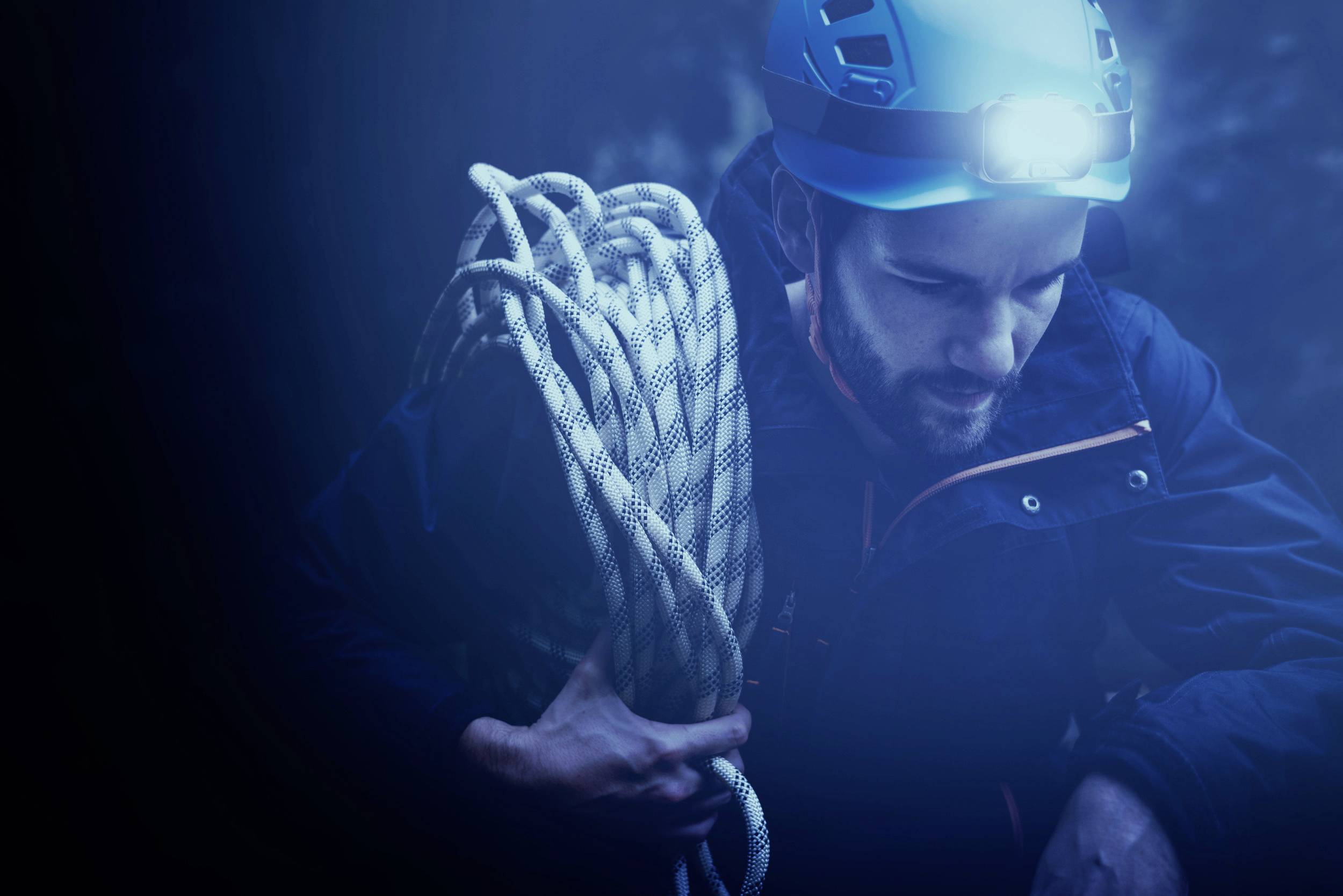 Ein Kletterer mit Helm hält ein Seil in einer dunklen Umgebung. Eine Stirnlampe leuchtet seinen Weg.