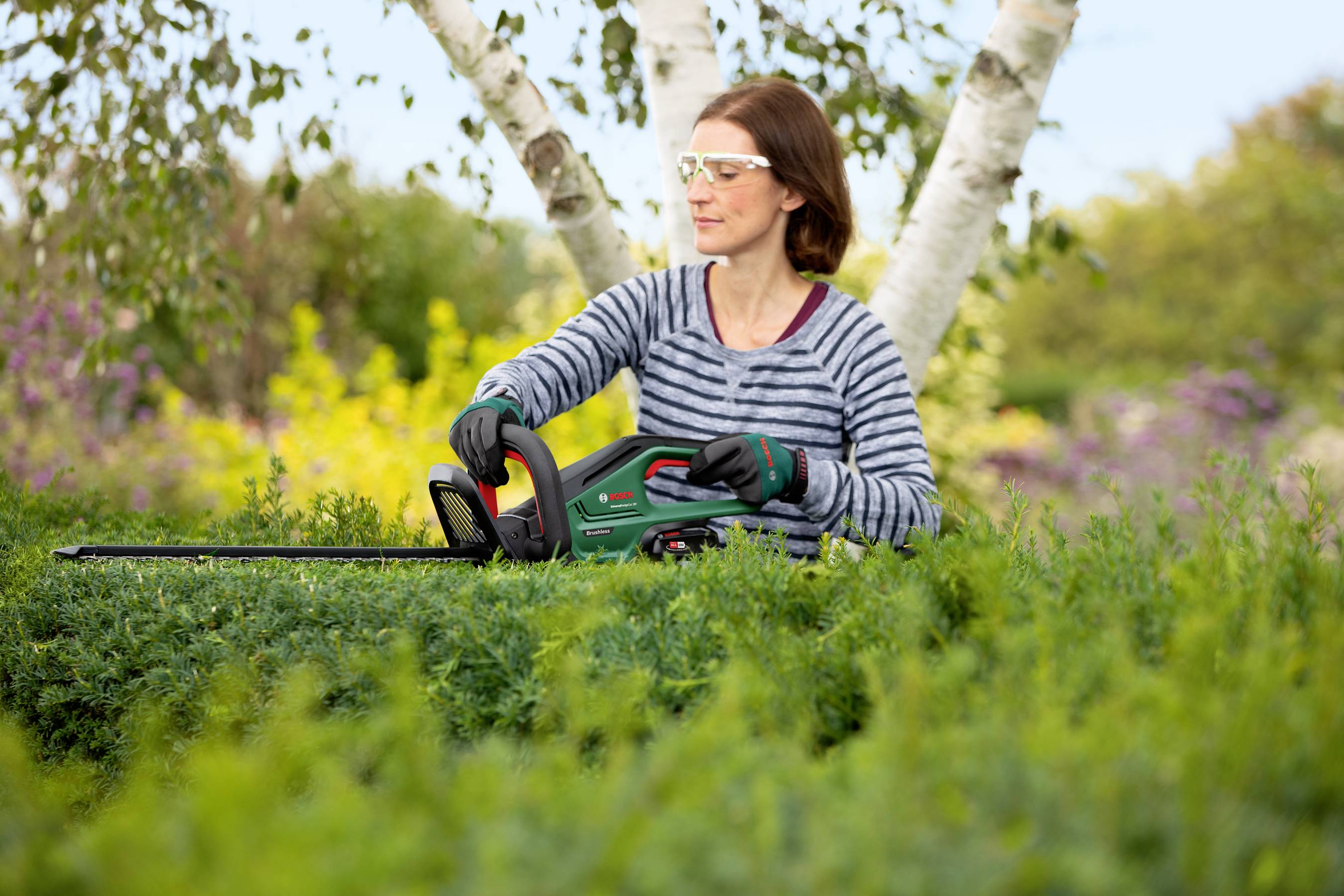 Eine Person in Schutzbrille und Handschuhen schneidet mit einer elektrischen Heckenschere eine Hecke, umgeben von Bäumen und Pflanzen.
