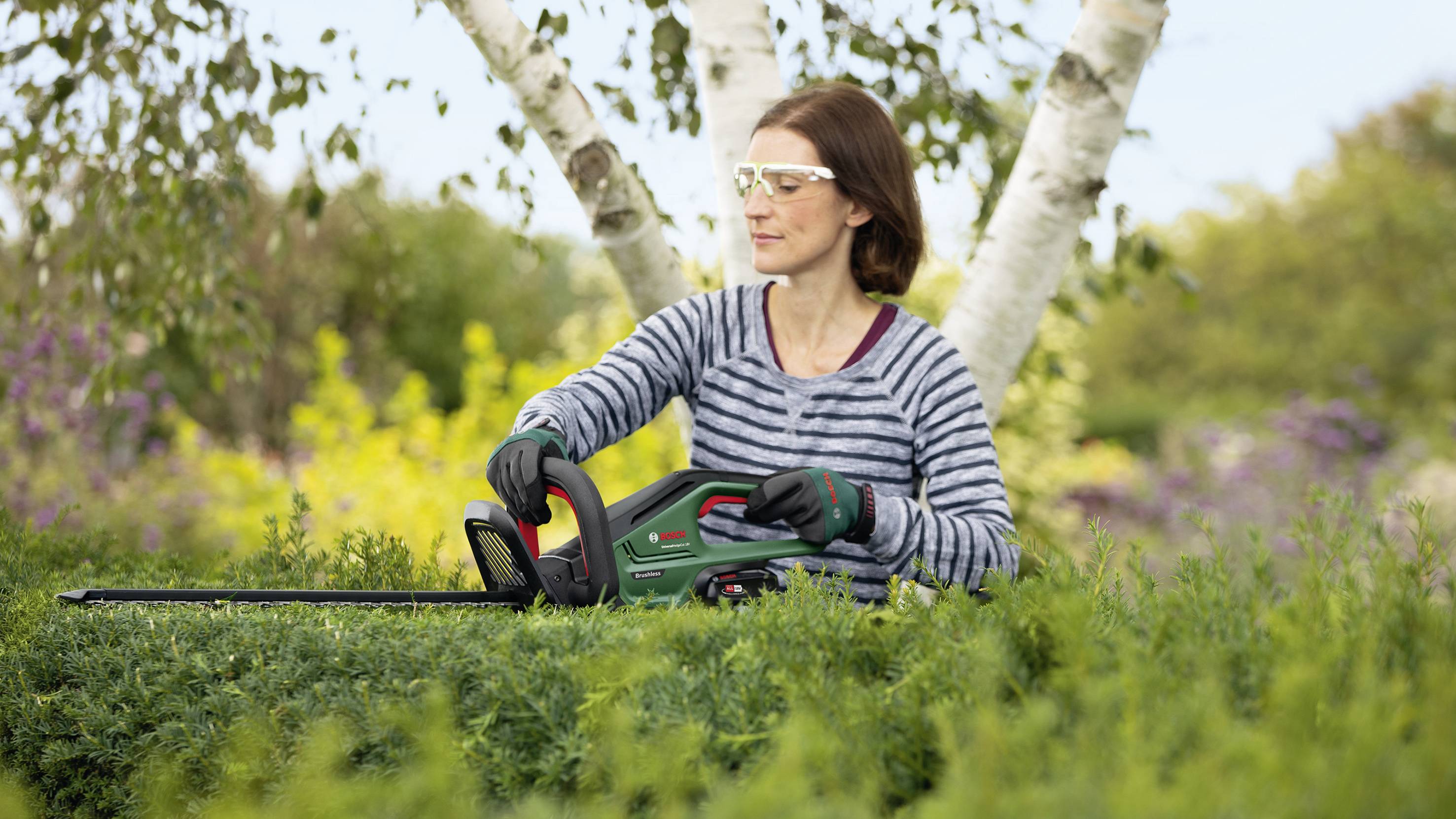 Eine Person schneidet mit einer elektrischen Heckenschere eine Hecke. Im Hintergrund sind Bäume und blühende Pflanzen zu sehen.