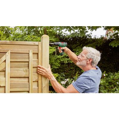 Ein älterer Mann montiert ein Holzpaneel mit einem Akkuschrauber im Garten, umgeben von grünen Bäumen und Sträuchern.