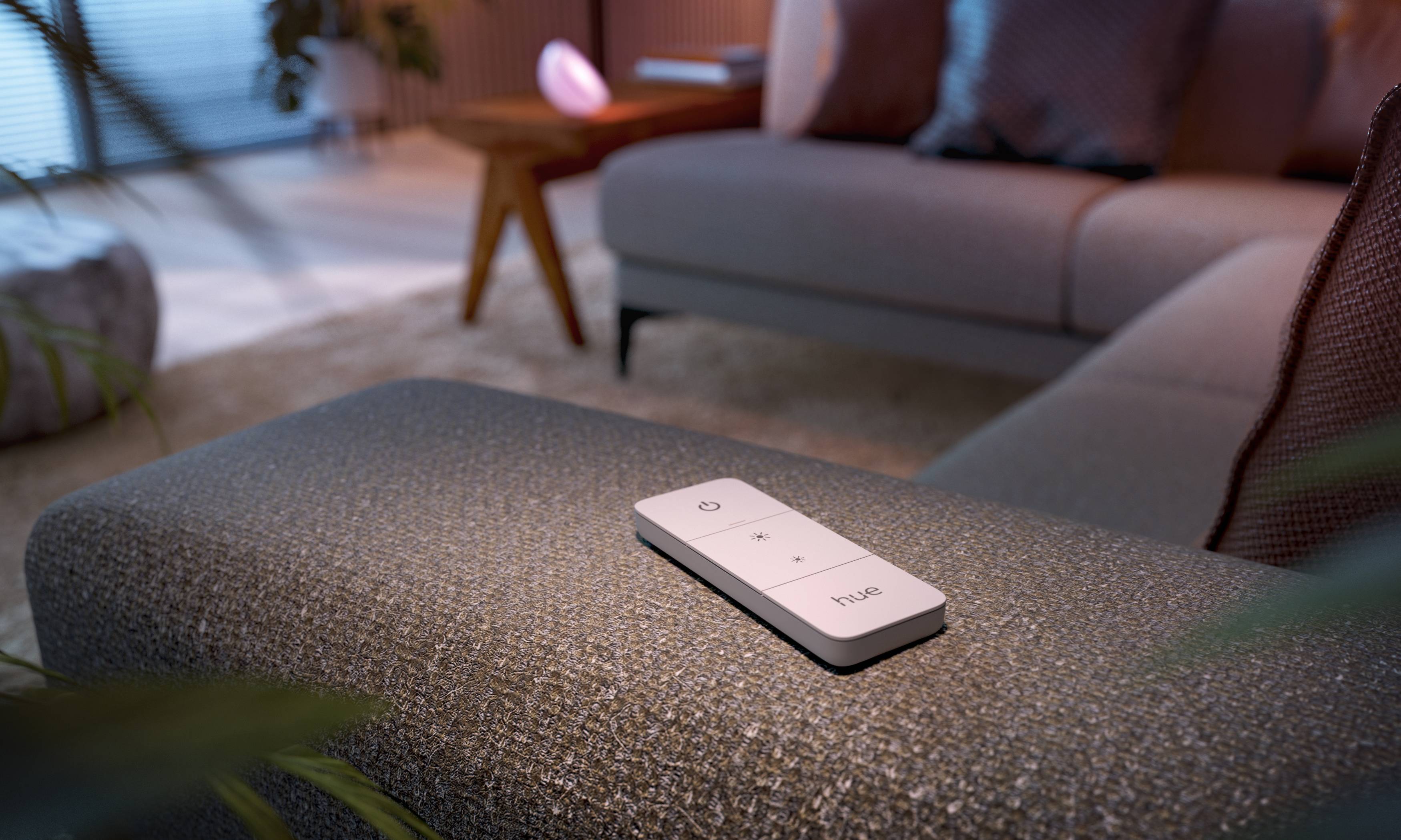 Fernbedienung auf grauem Sofa im modernen Wohnzimmer. Im Hintergrund dezente Beleuchtung und Holztisch, gemütliche Atmosphäre.