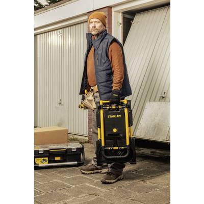 Ein Mann steht vor einer Garage und hält einen zusammengeklappten Wagen mit der Aufschrift 'Stanley'. Nebendran steht ein Werkzeugkoffer.