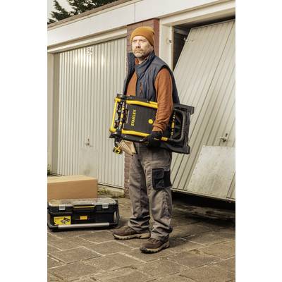 Ein Mann mit Mütze und Handschuhen steht vor einer Garage und hält ein gelbes Werkzeug, umgeben von einer Werkzeugkiste und einem Karton.