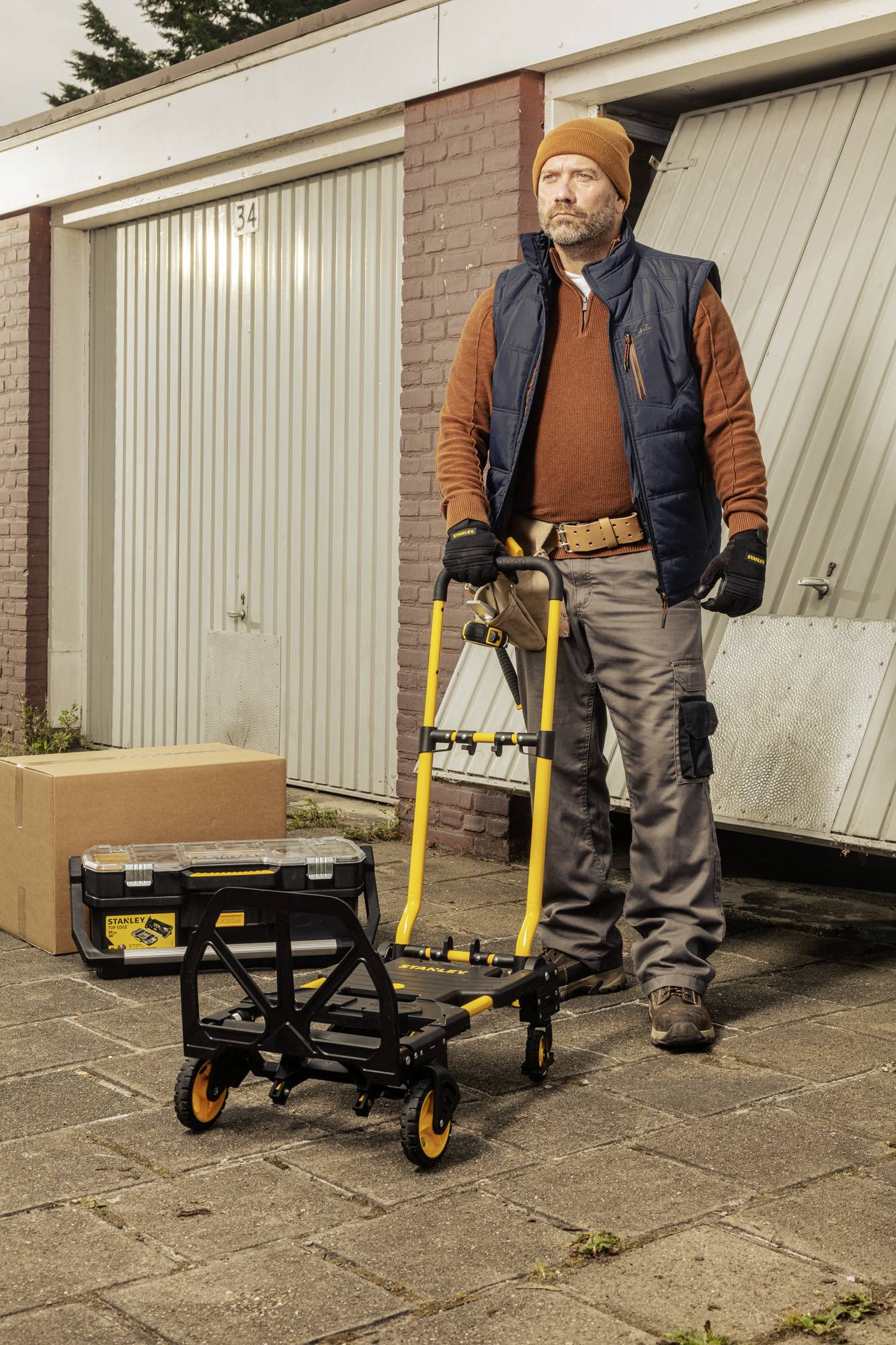 Ein Mann in Arbeitskleidung steht mit einem Handwagen vor einer offenen Garagentür. Neben ihm liegen eine Kiste und ein Karton.