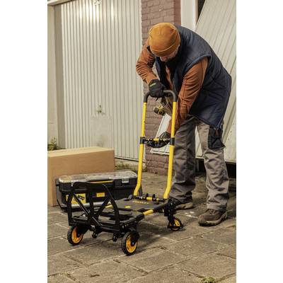 Eine Person schiebt einen gelben Handwagen, beladen mit einem Werkzeugkasten, auf einem gepflasterten Hof vor einer Garage.
