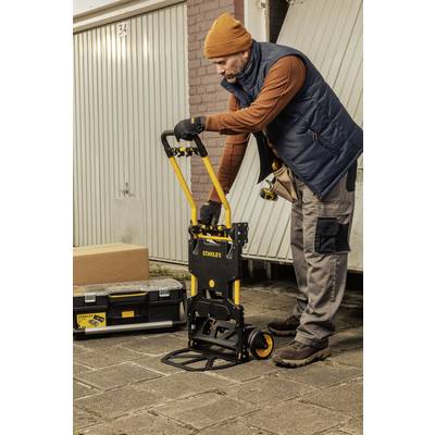 Ein Mann mit blauer Weste und orangefarbener Mütze benutzt einen schwarzen Handwagen, um eine Kiste in einer Garageneinfahrt zu transportieren.