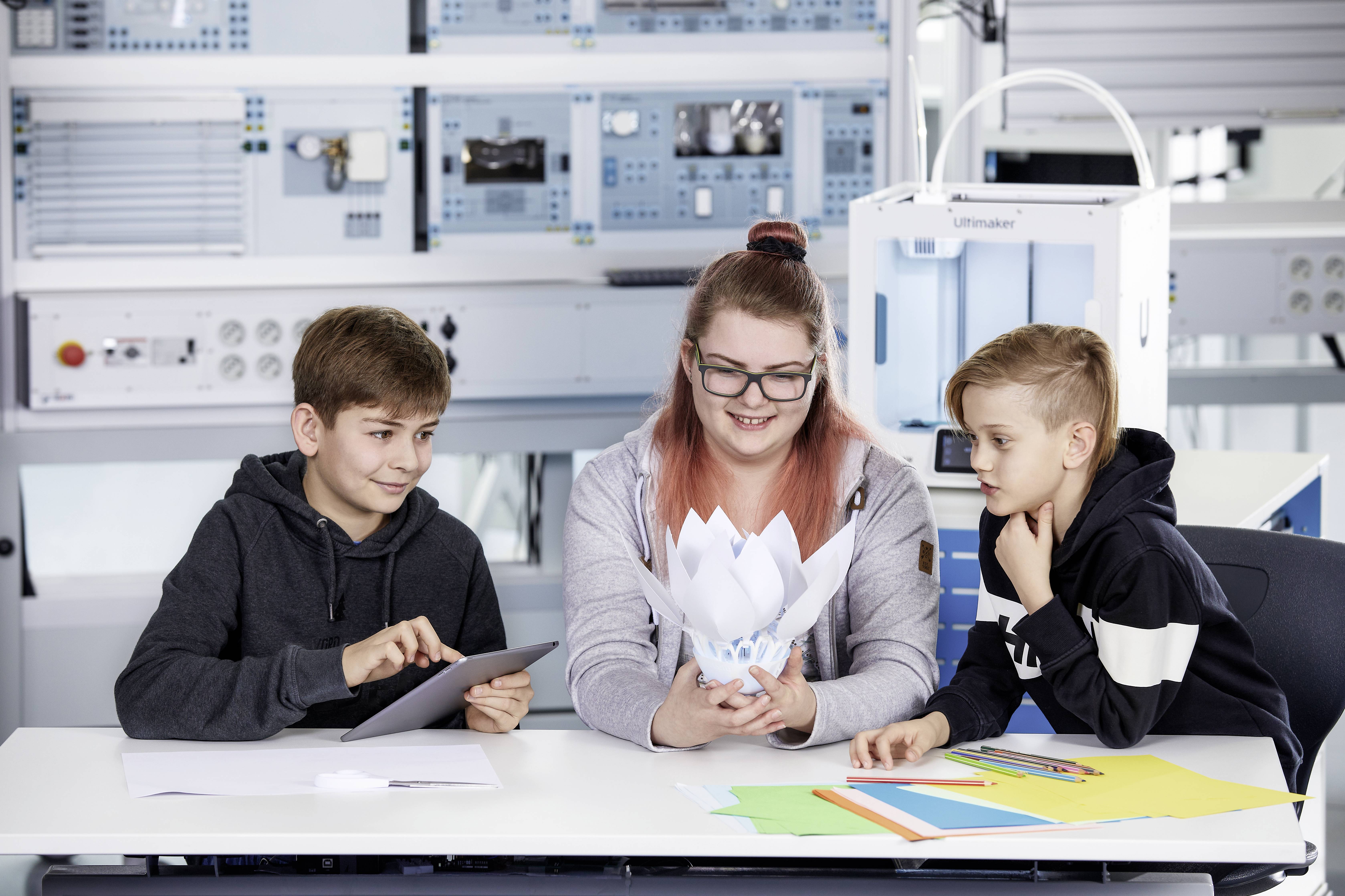Drei Kinder im Klassenzimmer mit einem Tablet und Papierprojekten. Sie sind an einem Tisch und scheinen an einem gemeinsamen Projekt zu arbeiten. Technische Geräte im Hintergrund.