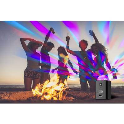 Eine Gruppe von Menschen tanzt fröhlich bei Sonnenuntergang um ein Lagerfeuer am Strand. Bunte Lichter erhellen die Szene.