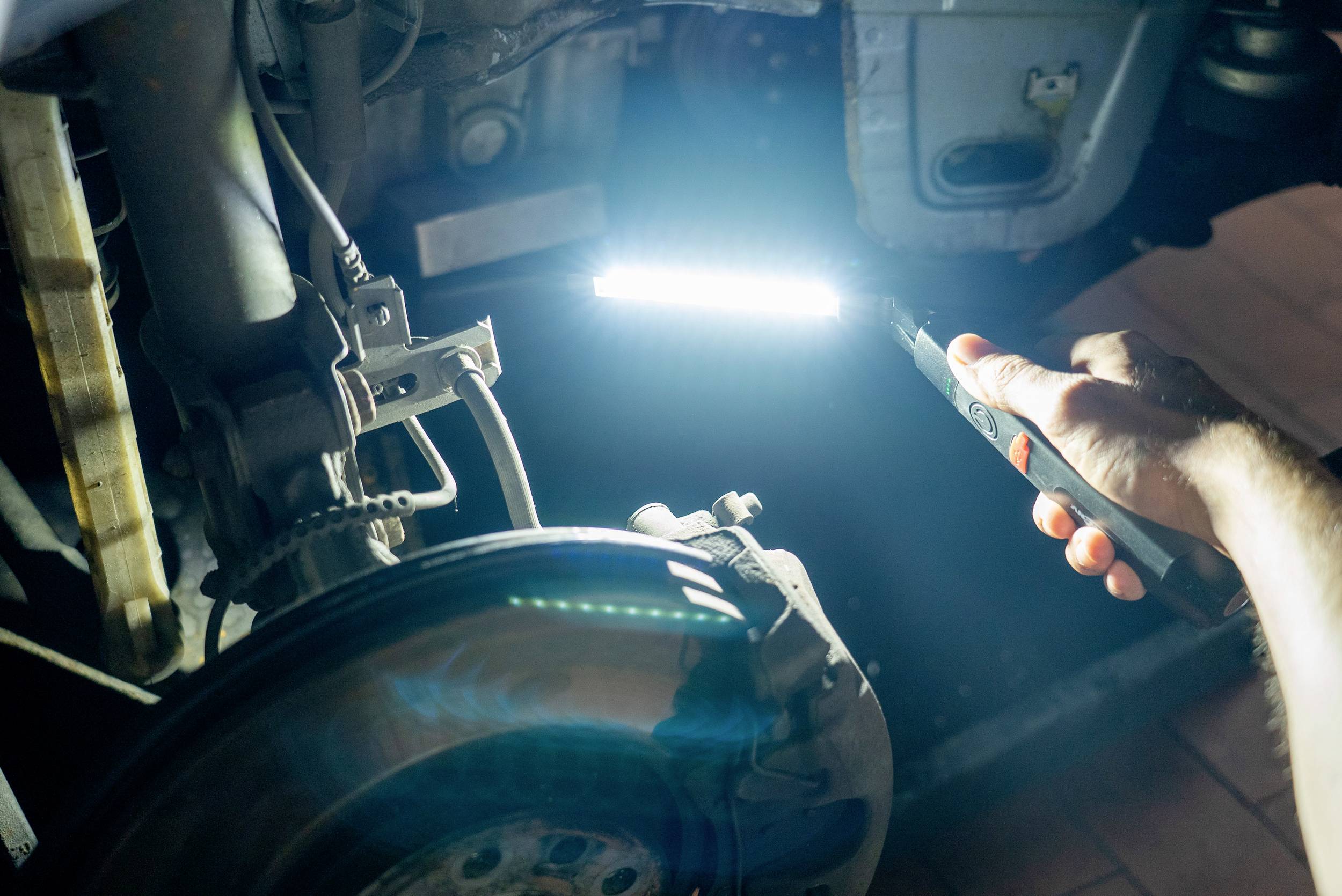 Eine Person hält eine Taschenlampe und leuchtet auf eine Autoachse oder ein Fahrzeugteil. Es wird in einer Werkstatt untersucht.