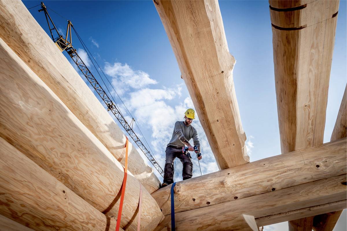 Ein Bauarbeiter in Schutzkleidung arbeitet hoch oben an einer Holzkonstruktion. Im Hintergrund ist ein Kran vor blauem Himmel zu sehen.