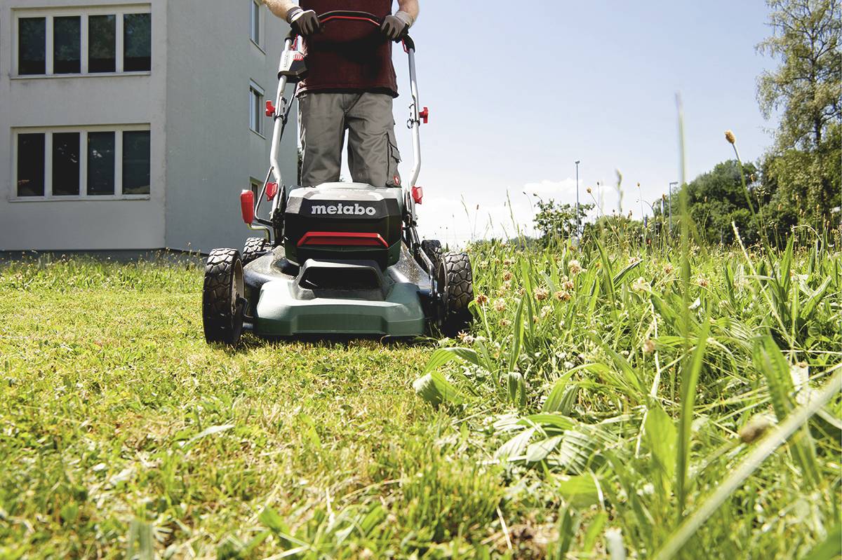 Eine Person mäht Rasen mit einem Metabo-Rasenmäher vor einem Gebäude, umgeben von Bäumen und Wiese an einem sonnigen Tag.