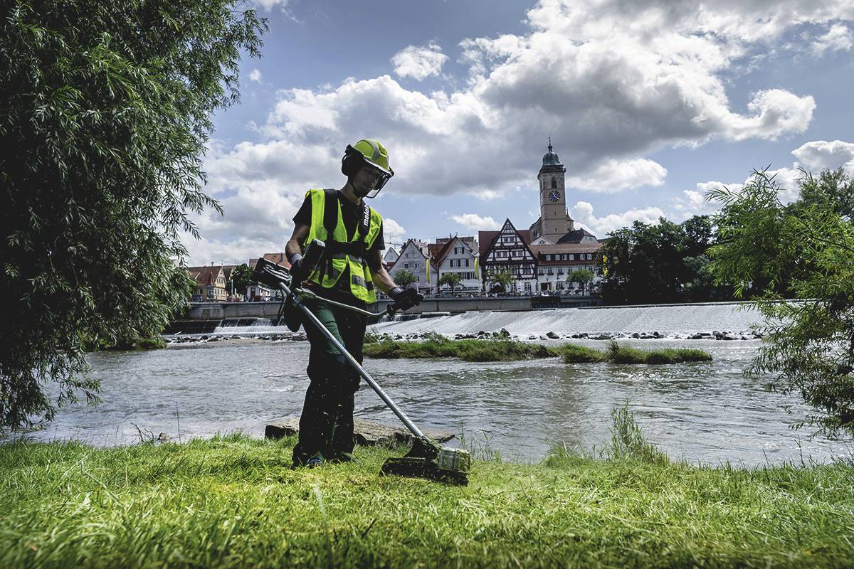 Eine Person in Sicherheitskleidung mäht mit einem Trimmer Rasen am Fluss. Im Hintergrund sind eine Stadt und eine Kirche sichtbar.