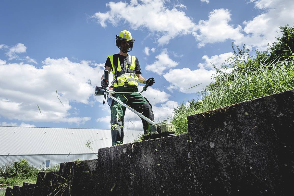 Eine Person in Schutzkleidung schneidet mit einem Rasentrimmer Gras auf betonierten Stufen unter einem blau bewölkten Himmel.