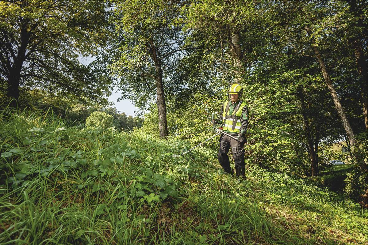Eine Person mit Schutzkleidung und Helm schneidet Gras und Unkraut mit einem Freischneider in einem bewaldeten Gebiet.