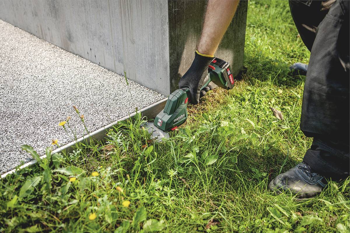 Eine Person schneidet mit einem elektrischen Rasentrimmer das Gras entlang der Kante eines Gebäudes. Sie trägt schwarze Handschuhe.