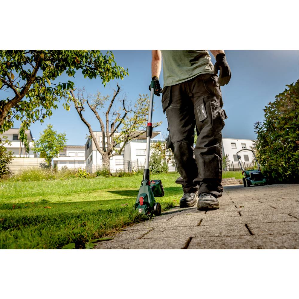 Eine Person mit Handschuhen schneidet den Rand eines Rasenplatzes an einem sonnigen Tag mit einem Gartengerät, wobei Häuser und Bäume im Hintergrund zu sehen sind.