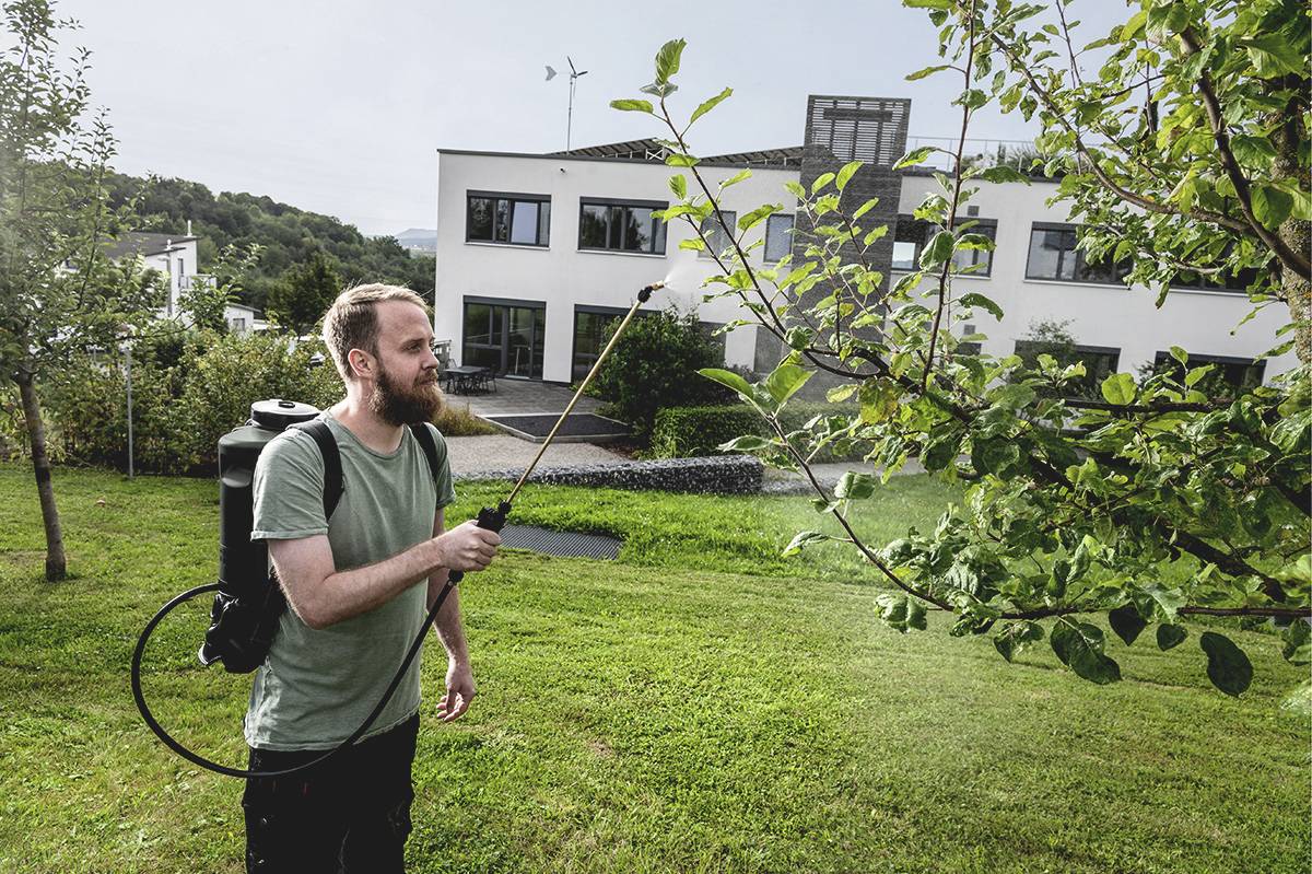 Ein Mann sprüht Flüssigkeit auf einen Baum in einem Garten. Im Hintergrund ist ein modernes weißes Gebäude zu sehen.