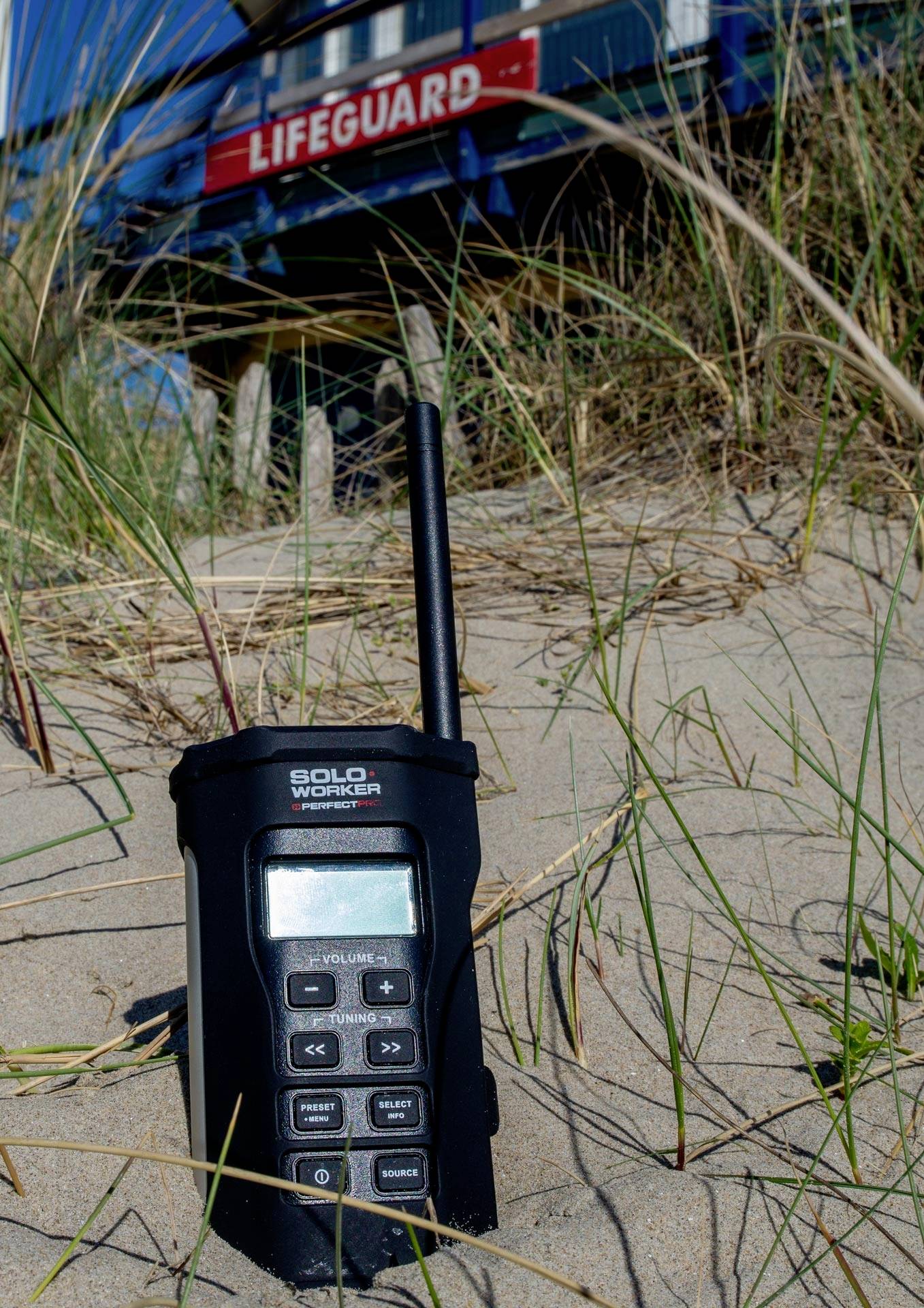 Funkgerät im Sand vor einem Rettungsschwimmer-Turm mit 'Lifeguard'-Schild. Gräser umgeben das Gerät.
